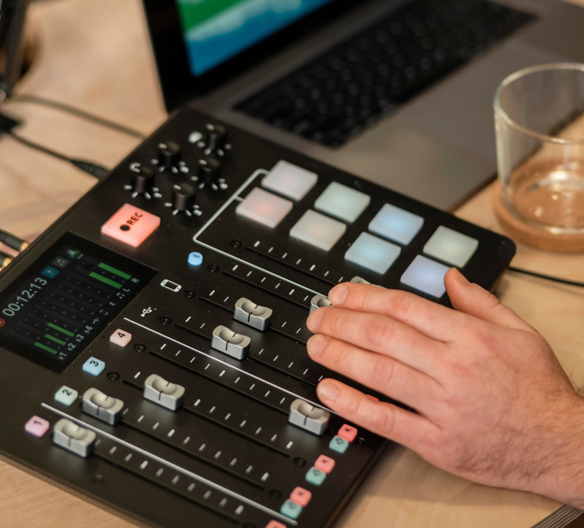 Person using a music production controller with a laptop displaying audio waves on a wooden desk.